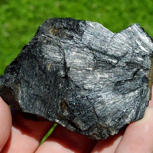 May include: A large, dark gray, rough-textured rock with a silvery sheen. The rock has a layered, striated appearance.