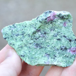 May include: A rough, green and black rock with small, pink inclusions. The rock is a type of gemstone called zoisite.