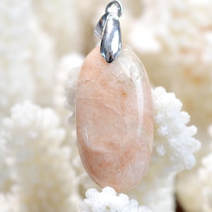 Puede incluir: Un colgante de piedra preciosa ovalado de color rosa pálido con una anilla de plata. El colgante está colocado sobre un fondo blanco.