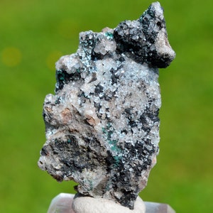 May include: A grey and black rock with small, white crystals and green accents. The rock has a rough, textured surface.