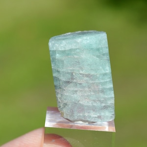 Puede incluir: Un cristal de aguamarina azul claro con estrías en la superficie. El cristal está en un soporte de exhibición transparente.