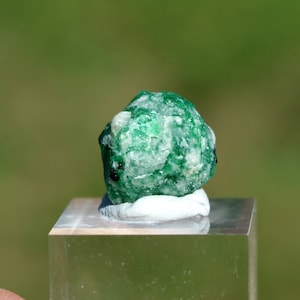 May include: A green emerald crystal with a rough, textured surface. The crystal is sitting on a white pedestal.