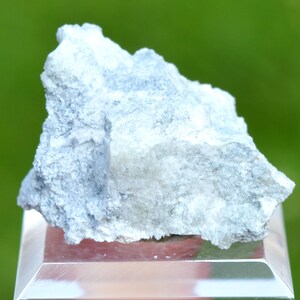 May include: A light blue mineral specimen with a rough, textured surface. The stone is displayed on a clear, rectangular stand, set against a blurred green background. The mineral's color ranges from pale blue to white.