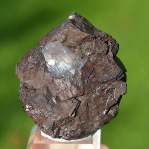 Puede incluir: Un espécimen mineral marrón oscuro y rugoso con un parche metálico plateado. La roca tiene una superficie irregular y dentada y se muestra sobre un fondo verde borroso. El mineral parece estar apoyado sobre un soporte transparente y angulado.