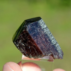 Puede incluir: Un cristal rojo oscuro facetado con una textura natural rugosa. El cristal está sentado en un soporte de exhibición transparente.