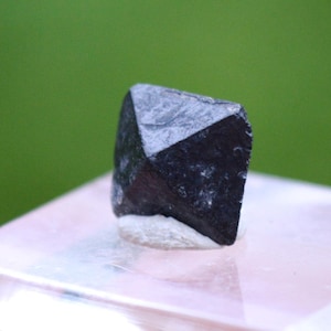 Op de afbeelding: Een donker, facetachtig, zwart mineraal specimen met een kristallijne structuur. De steen heeft een ruwe textuur en staat op een lichtgekleurde basis, met een vage groene achtergrond. Het mineraal lijkt een natuurlijke, ongepolijste steen te zijn.