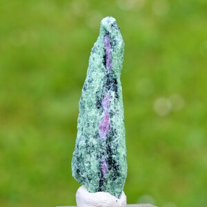 Peut inclure: Un cristal de zoïsite vert et rose avec une surface rugueuse et texturée. Le cristal est debout sur une base blanche.