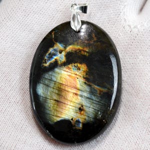Puede incluir: Colgante ovalado de labradorita con fianza de plata. La piedra pulida muestra un color base oscuro con destellos iridiscentes de azul, amarillo y naranja. La gema mide aproximadamente 3,8 cm de largo y 2,5 cm de ancho, mostrando un patrón natural único.