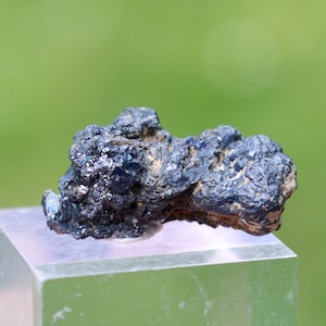 May include: A dark blue mineral specimen with a rough, textured surface. The mineral sits on a clear, rectangular display stand against a blurred green background. The specimen has a crystalline appearance.