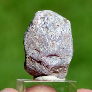 Könnte beinhalten: Ein unregelmäßig geformter Felsen mit einer Mischung aus Grau-, Lila- und Brauntönen. Die Oberfläche hat ein strukturiertes, geschichtetes Aussehen. Der Felsen wird auf einem transparenten, quadratischen Sockel vor einem verschwommenen grünen Hintergrund präsentiert.