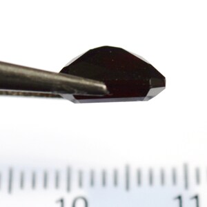May include: A dark red gemstone with a faceted cut, held by tweezers over a ruler marked with numbers 10 and 11.
