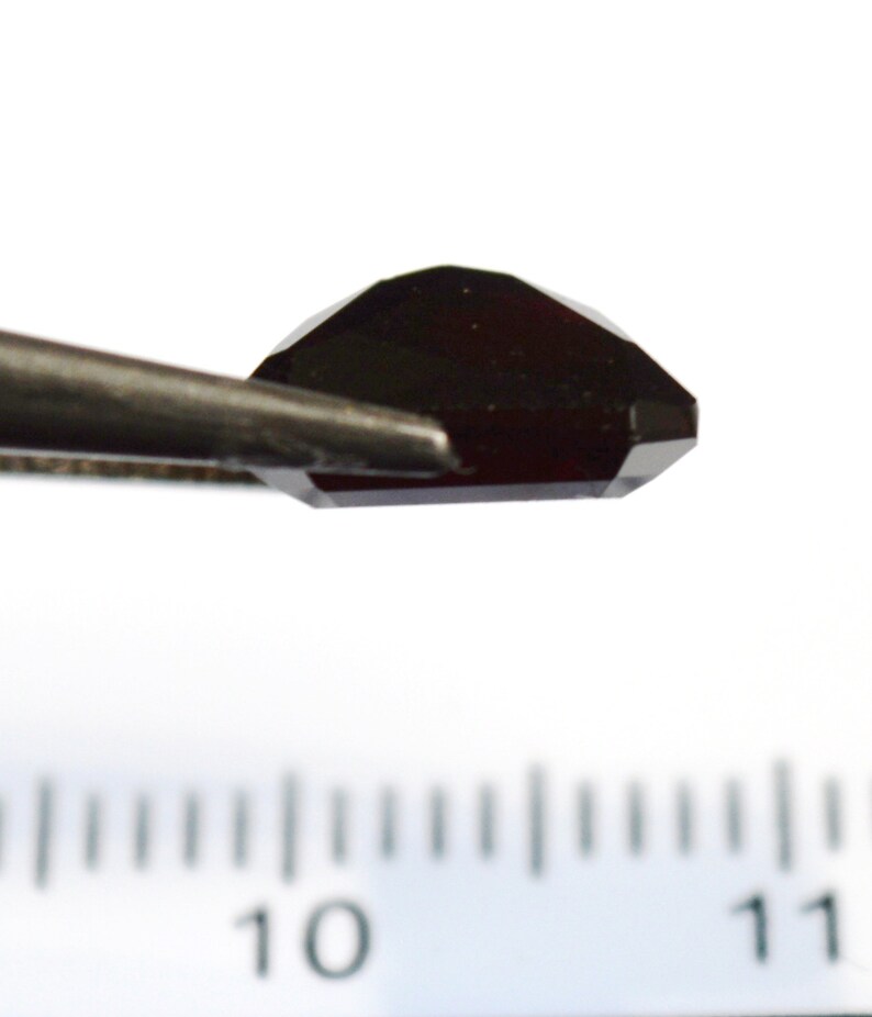 May include: A dark red gemstone with a faceted cut, held by tweezers over a ruler marked with numbers 10 and 11.