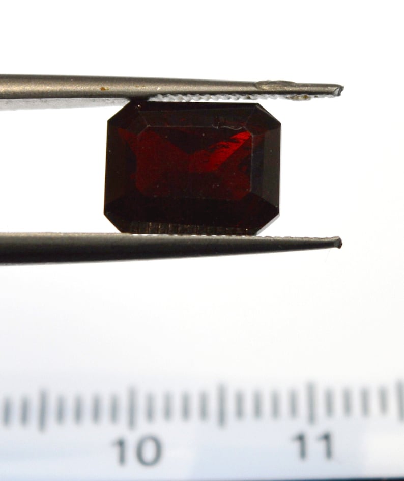 May include: A deep red gemstone, likely a garnet, is held by tweezers over a ruler marked with numbers 10 and 11. The gemstone is a rectangular emerald cut.