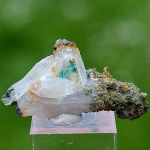 May include: A close-up of a mineral specimen featuring clear quartz crystals with green and brown inclusions. The specimen is mounted on a clear, rectangular stand against a blurred green background. The mineral has a rough, textured appearance.