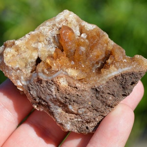 Könnte beinhalten: Ein brauner Geode mit einem Cluster aus orangefarbenen Calcitkristallen. Die Kristalle sind durchscheinend und haben einen glasartigen Glanz. Der Geode hat einen Durchmesser von etwa 7,5 cm.