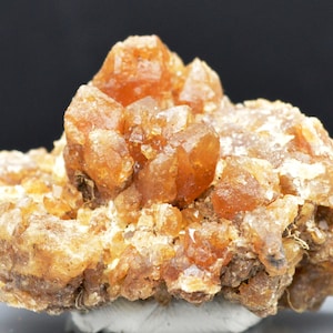 Könnte beinhalten: Ein Cluster aus orangefarbenen und braunen Kristallen auf einer rauen, braunen Matrix. Die Kristalle sind durchscheinend und haben einen glasartigen Glanz.