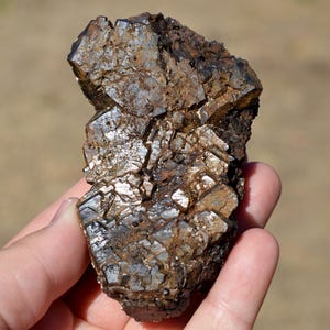 Könnte beinhalten: Ein rohes Mineralexemplar mit einer Ansammlung dunkelbrauner, metallischer und blockartiger Kristalle. Die Oberfläche weist eine komplexe, zerklüftete Textur mit reflektierenden Facetten und tiefen Erdtönen auf, die eine natürliche, ungeformte geologische Struktur zeigt.