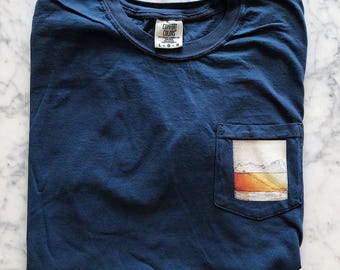 May include: A navy blue t-shirt with a pocket on the left side. The pocket has a graphic of a sunset with orange, yellow, and red colors. The t-shirt has a tag that says "Comfort Colors" and "L-O-G".