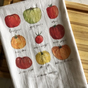 May include: White kitchen towel featuring watercolor illustrations of various heirloom tomatoes, including Brandywine, Green Giant, and Kellogg's Breakfast. The towel has the words "HEIRLOOM TOMATOES" printed at the bottom.