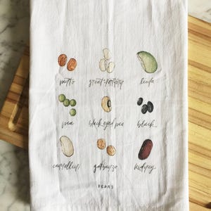 May include: White kitchen towel with watercolor illustrations of various beans, including pinto, great northern, and kidney beans. Each bean type is labeled with its name in a handwritten font. The word "BEANS" is written at the bottom.