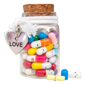 Pode incluir: Um frasco de vidro cheio de c&aacute;psulas coloridas com carinhas felizes. O frasco tem uma tampa de corti&ccedil;a e um cord&atilde;o branco amarrado ao redor. Um amuleto de cora&ccedil;&atilde;o prateado com a palavra "LOVE" est&aacute; amarrado ao cord&atilde;o. Uma c&aacute;psula est&aacute; fora do frasco, com um bilhete amarelo dentro.