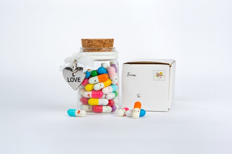 Pode incluir: Um frasco de vidro cheio de c&aacute;psulas coloridas com carinhas sorridentes, um amuleto de cora&ccedil;&atilde;o prateado com a palavra "LOVE" e uma caixa branca com as palavras "De" e "Para" escritas nela.