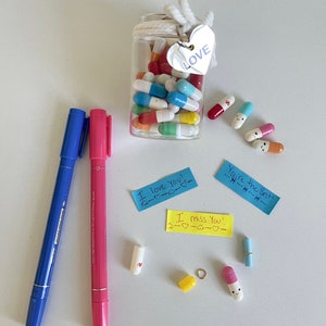 Pode incluir: Um frasco de vidro cheio de c&aacute;psulas coloridas com mensagens escritas &agrave; m&atilde;o dentro. O frasco tem um cord&atilde;o branco amarrado ao redor com uma etiqueta de cora&ccedil;&atilde;o prateada que diz "LOVE".  Existem v&aacute;rias c&aacute;psulas espalhadas ao redor do frasco com notas escritas &agrave; m&atilde;o que dizem "I love you!" e "I miss you!"