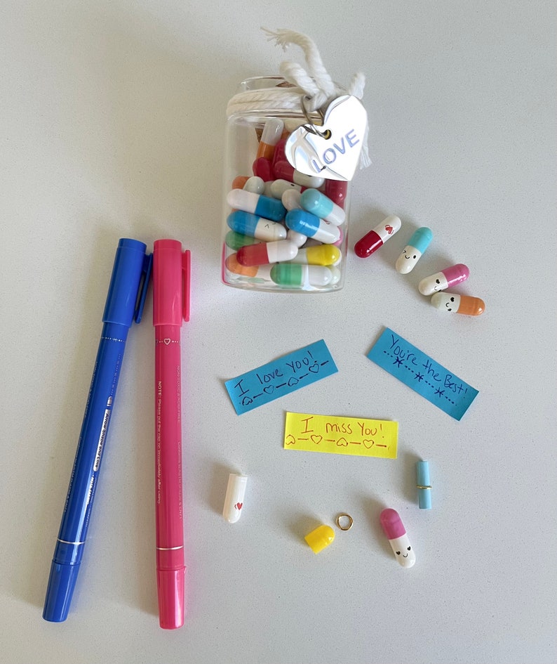 Pode incluir: Um frasco de vidro cheio de c&aacute;psulas coloridas com mensagens escritas &agrave; m&atilde;o dentro. O frasco tem um cord&atilde;o branco amarrado ao redor com uma etiqueta de cora&ccedil;&atilde;o prateada que diz "LOVE".  Existem v&aacute;rias c&aacute;psulas espalhadas ao redor do frasco com notas escritas &agrave; m&atilde;o que dizem "I love you!" e "I miss you!"