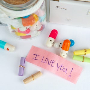 Pode incluir: Um frasco de vidro cheio de c&aacute;psulas coloridas com carinhas sorridentes, um bilhete rosa que diz "Eu te amo!" e v&aacute;rias notas enroladas amarradas com um cord&atilde;o dourado.
