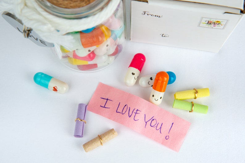 Pode incluir: Um frasco de vidro cheio de c&aacute;psulas coloridas com carinhas sorridentes, um bilhete rosa que diz "Eu te amo!" e v&aacute;rias notas enroladas amarradas com um cord&atilde;o dourado.