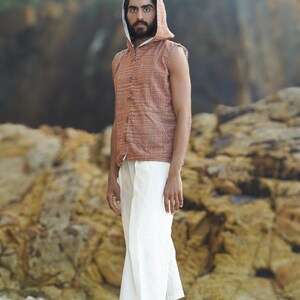 May include: A man wearing a sleeveless, rust-colored, button-up vest with a hood. The vest has a white trim around the hood and neckline. He is wearing white pants and is standing on a sandy beach.