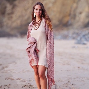 Op de afbeelding: Een vrouw die een lichtbruine jurk draagt met een roze en rode sjaal. Ze staat op een zandstrand met een klif op de achtergrond.