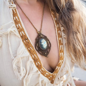 May include: A handmade macrame necklace with a large, oval pendant. The pendant has a blue and green stone set in a brown macrame design. The necklace is worn over a cream-coloured top with brown trim.