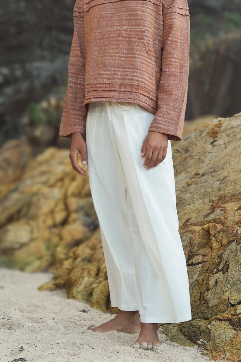 May include: A person wearing a rust-colored striped top with a white button closure and white wide-leg pants.
