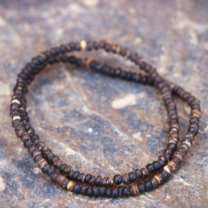 May include: Two strands of dark brown beaded necklace, with small, round beads. Some beads have lighter brown accents. The necklace is displayed on a textured, neutral-toned surface, creating a natural, earthy aesthetic.