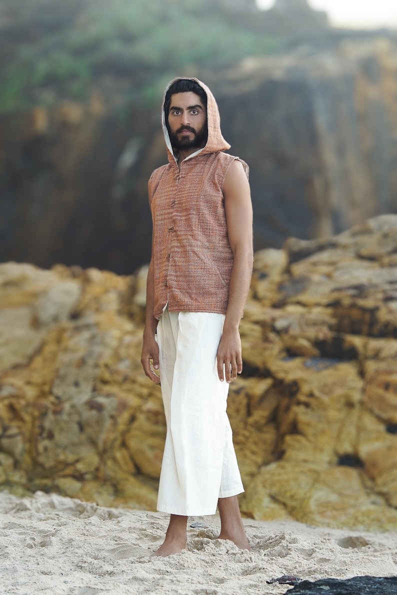 May include: A man wearing a sleeveless, rust-colored, button-up vest with a hood. The vest has a white trim around the hood and neckline. He is wearing white pants and is standing on a sandy beach.