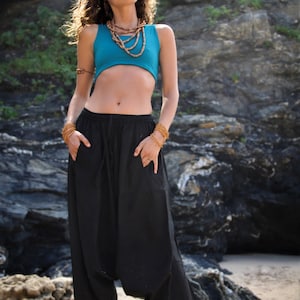 May include: A woman wearing a turquoise crop top and black harem pants. She is standing on a sandy beach with a rocky cliff in the background.