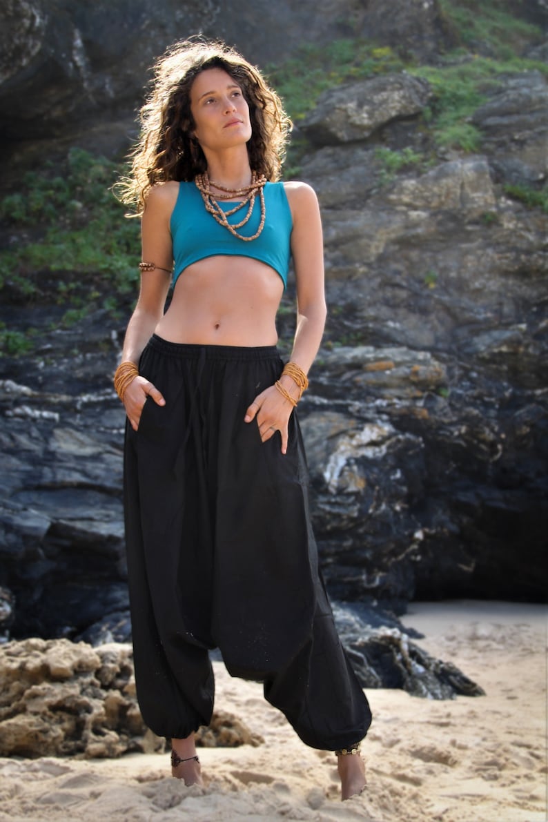 May include: A woman wearing a turquoise crop top and black harem pants. She is standing on a sandy beach with a rocky cliff in the background.