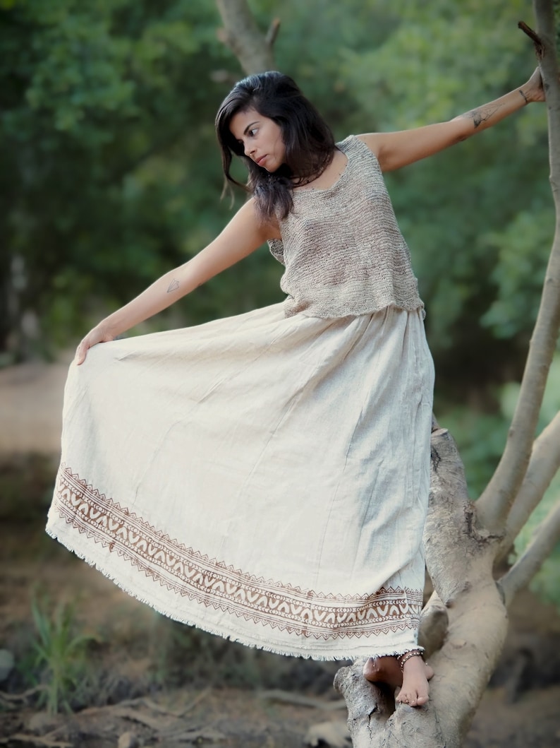 May include: A woman wearing a white skirt with a brown patterned border and a brown sleeveless top. She is standing on a tree branch in a forest setting.
