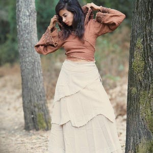May include: A woman wearing a brown hooded top with a white tiered skirt. She is standing in a forest setting.