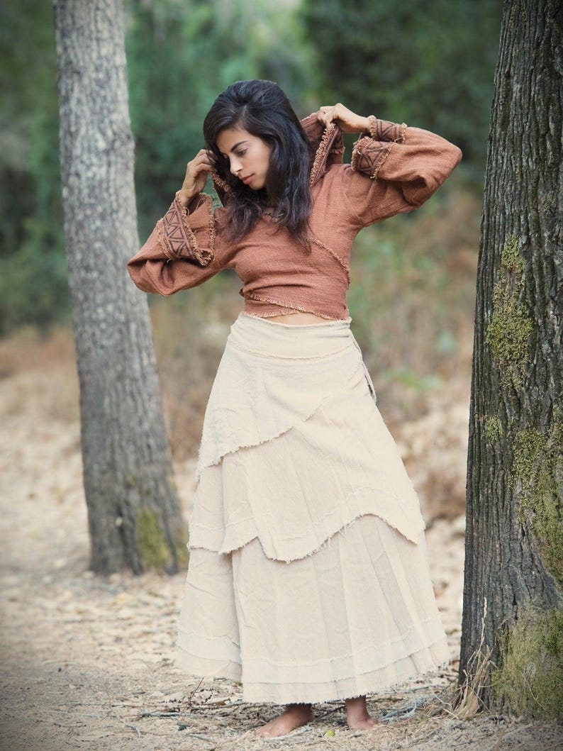 May include: A woman wearing a brown hooded top with a white tiered skirt. She is standing in a forest setting.
