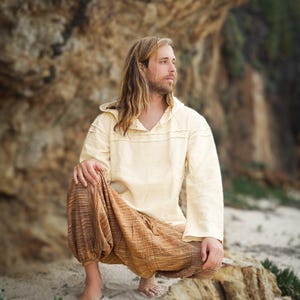 May include: A person wearing a cream-colored hooded shirt and brown striped pants sits on a rock with a sandy beach in the background.