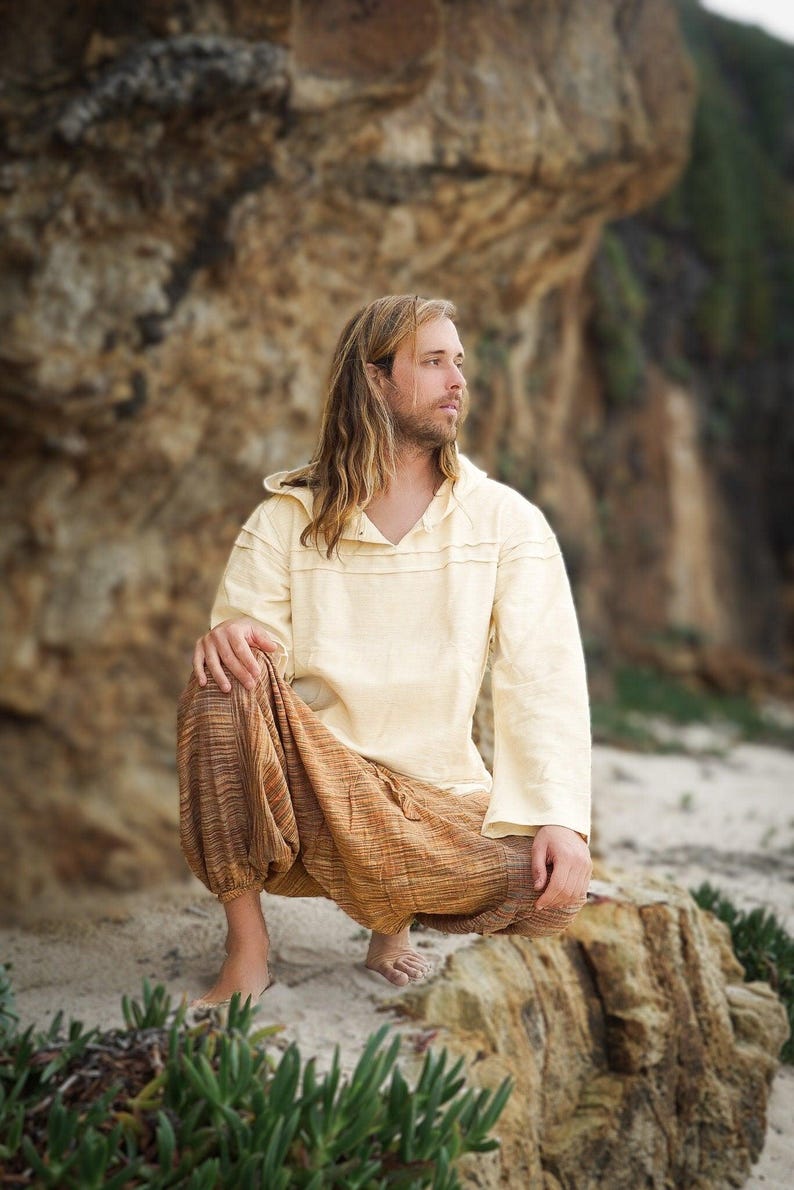 May include: A person wearing a cream-colored hooded shirt and brown striped pants sits on a rock with a sandy beach in the background.