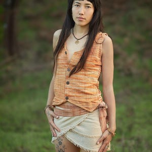 May include: A woman wearing a sleeveless, orange and white striped top with a brown button closure. She is also wearing a white wrap skirt with a brown belt and a gold necklace with a tree pendant. She has long dark hair and is standing in a grassy field.