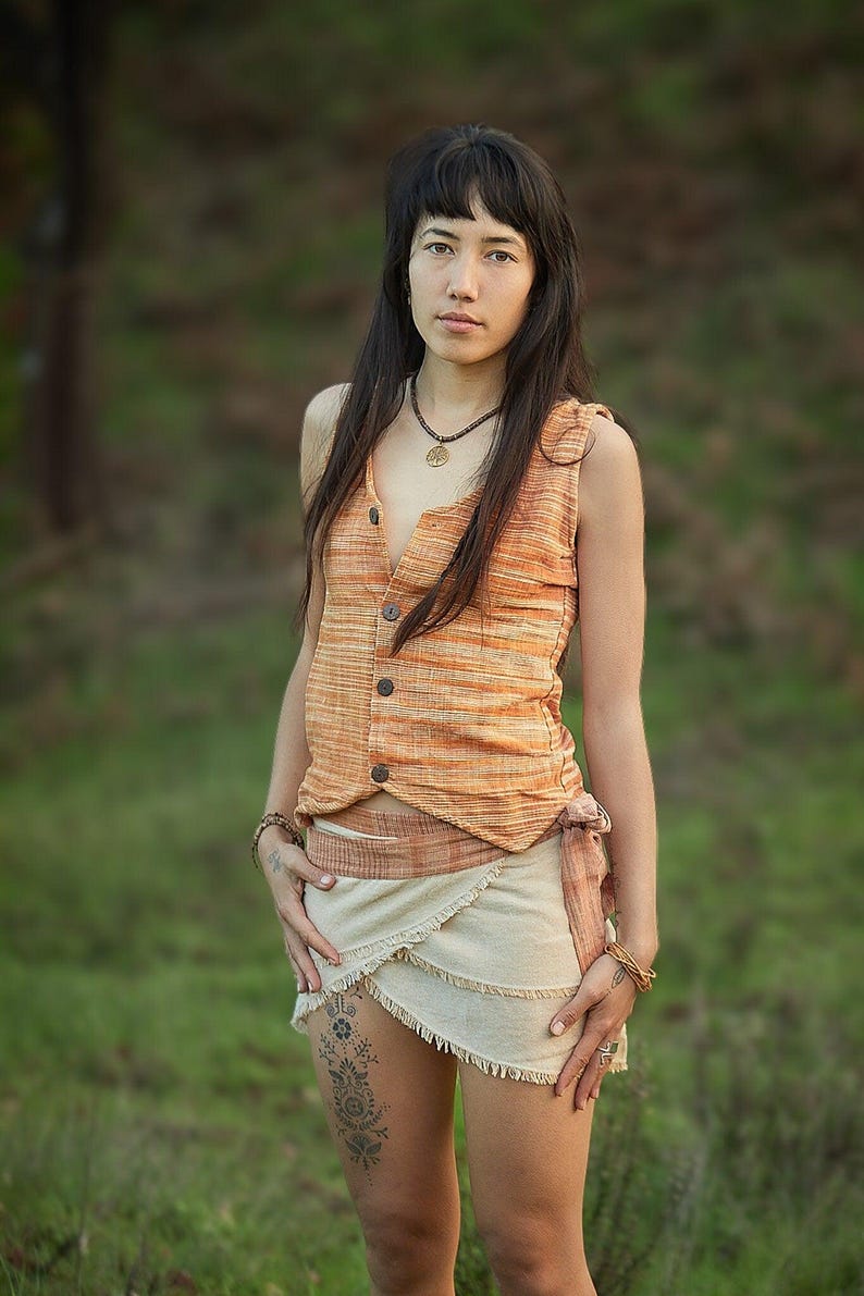 May include: A woman wearing a sleeveless, orange and white striped top with a brown button closure. She is also wearing a white wrap skirt with a brown belt and a gold necklace with a tree pendant. She has long dark hair and is standing in a grassy field.