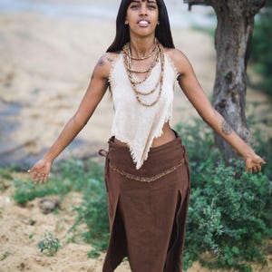 May include: A woman wearing a white, sleeveless top with a brown skirt and multiple beaded necklaces. She is standing in a desert landscape with sand and green plants.