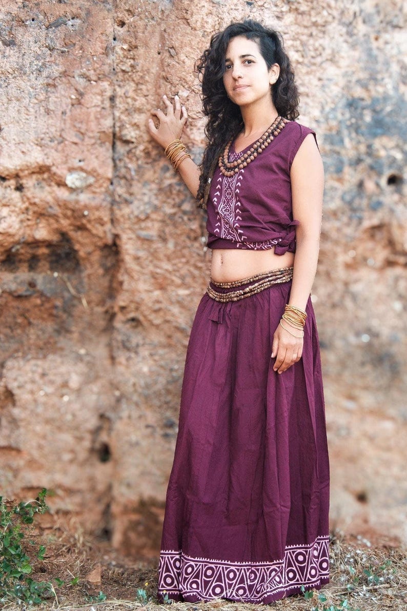 May include: A woman wearing a purple top with a white geometric pattern and a matching purple skirt with a white geometric border. She is wearing a brown beaded necklace and multiple gold bracelets.
