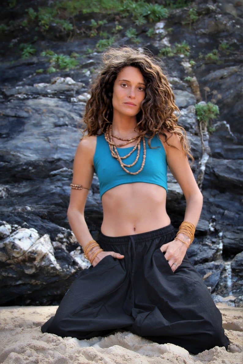 May include: A woman wearing a turquoise crop top and black pants with a wooden beaded necklace. She is kneeling on a sandy beach with a rocky cliff in the background.