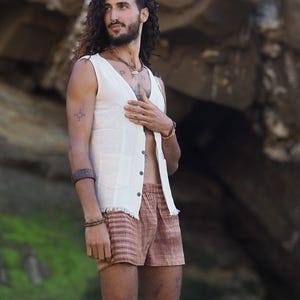 Puede incluir: Un hombre con un chaleco blanco sin mangas de lino con un patrón de rayas marrones y blancas en la parte inferior. Lleva un collar con un colgante y tiene un tatuaje en el brazo.