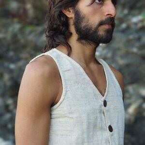 May include: A man with long dark hair and a beard is wearing a sleeveless, off-white linen vest with brown buttons. The vest has a V-neck and is made of a textured fabric.